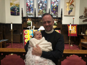 a smiling priest holds a baby in a baptismal gown with the church altar as a background