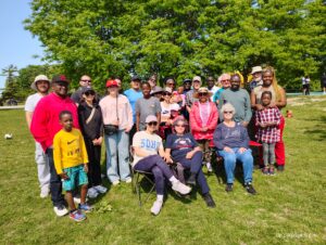 Group of diverse smiling people in casual clothing outdoors in the sunshine. Most people are standing, others are sitting. 