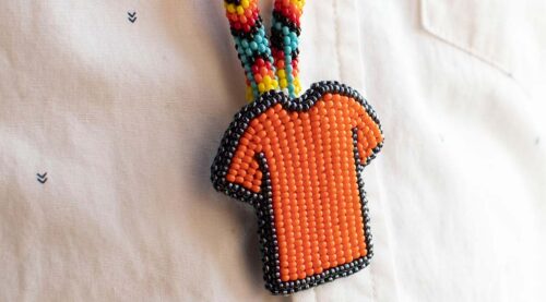 Depicts a beaded pendant in the shape of an orange shirt with a black border. The pendant is on a beaded chain in many colours